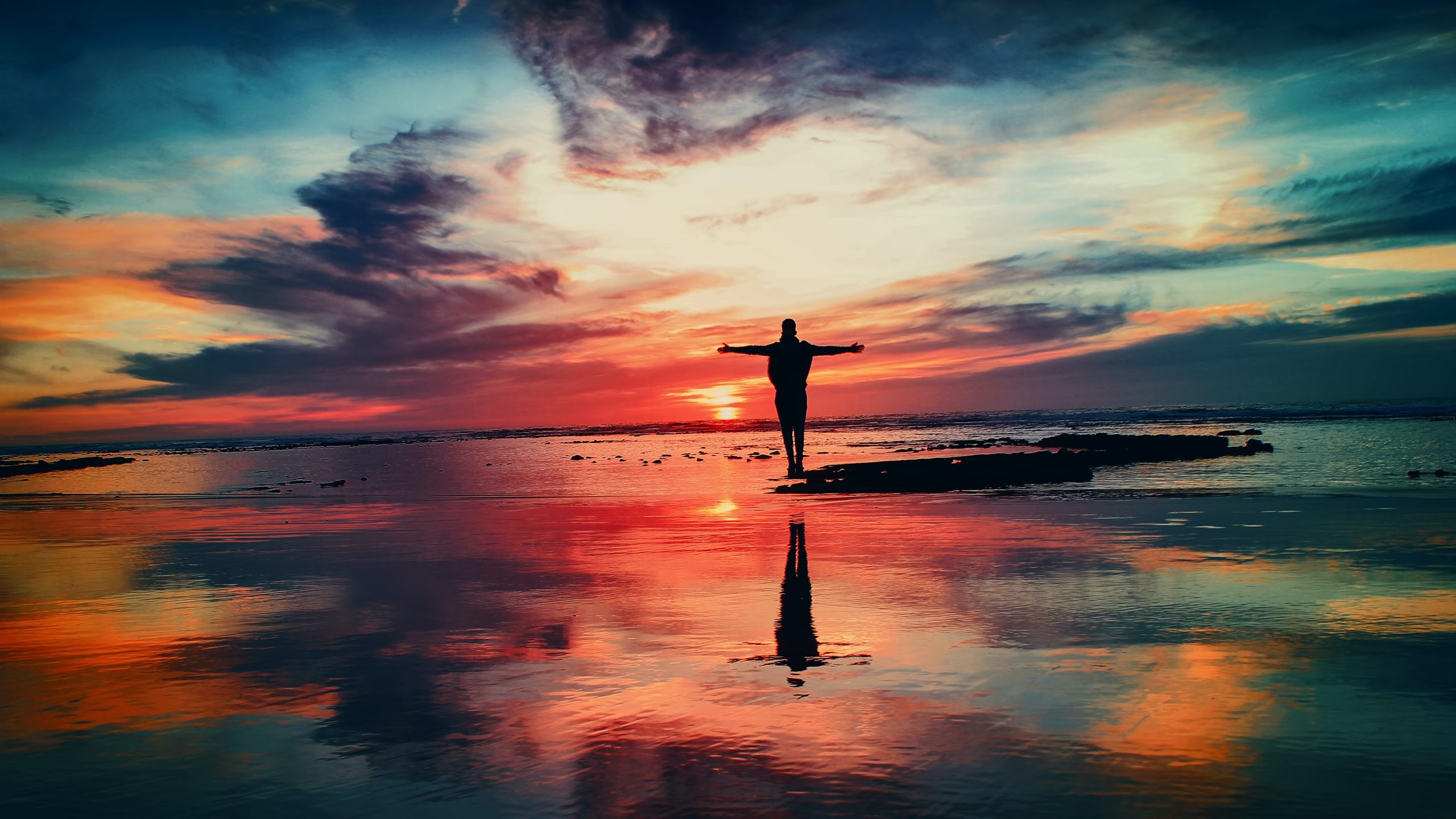 En person som står på en sten i vattnet och ser mot solnedgången i blå-gul-röda toner